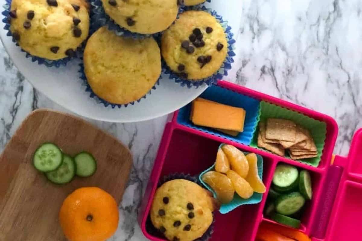A plate of orange creamsicle muffins can be seen in the top left of the image. The bottom right corner of the image contains a wooden cutting board that has three cucumber slices and a clementine on top of it. The bottom left corner contains a plastic lunch tray with several divided compartments. The compartments contain an orange creamsicle muffin, clementines, cucumbers, cheese, crackers, and bell peppers.