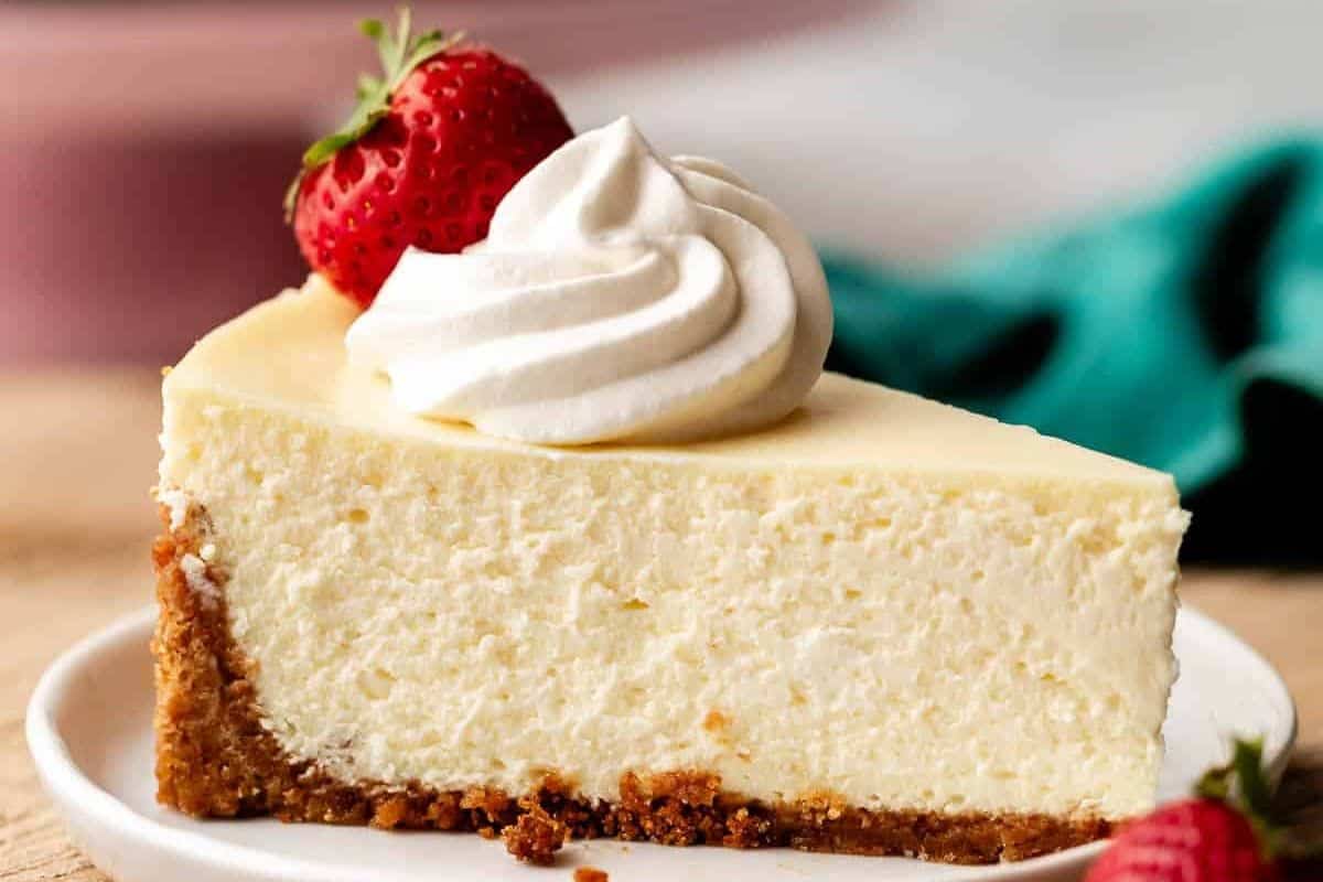 A slice of cheesecake topped with a dollop of whipped cream and garnished with a fresh strawberry on a white plate, this treat is the epitome of old-fashioned desserts.