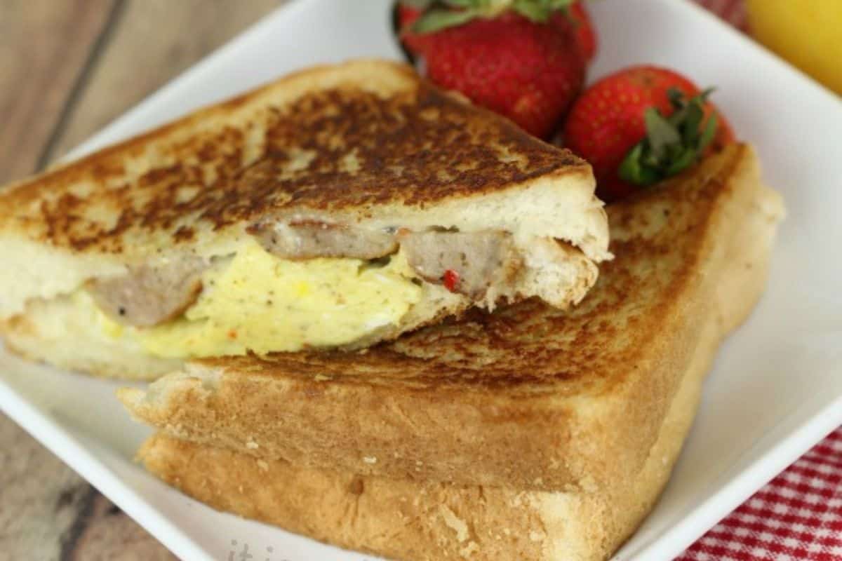 A grilled breakfast sandwich filled with scrambled eggs and sausage is cut in half and served on a white plate with three strawberries.