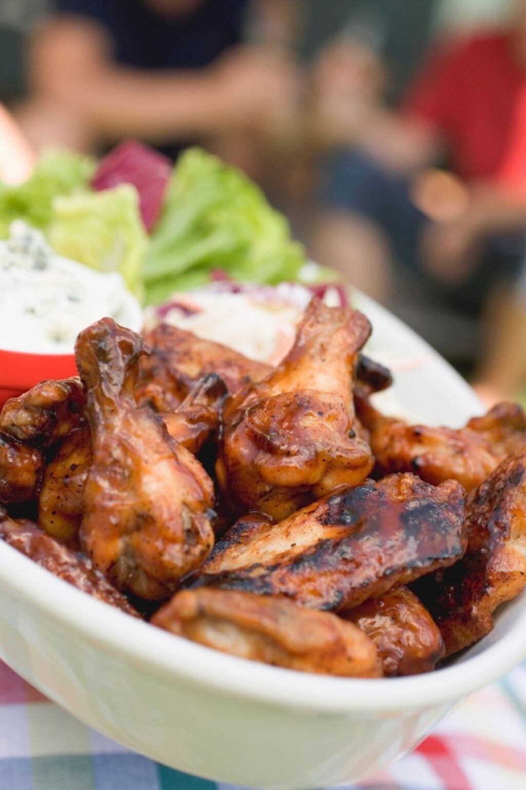 A white bowl filled with Grilled Chicken Wings sits on a table, accompanied by lettuce, dip, and other salad ingredients in the background.