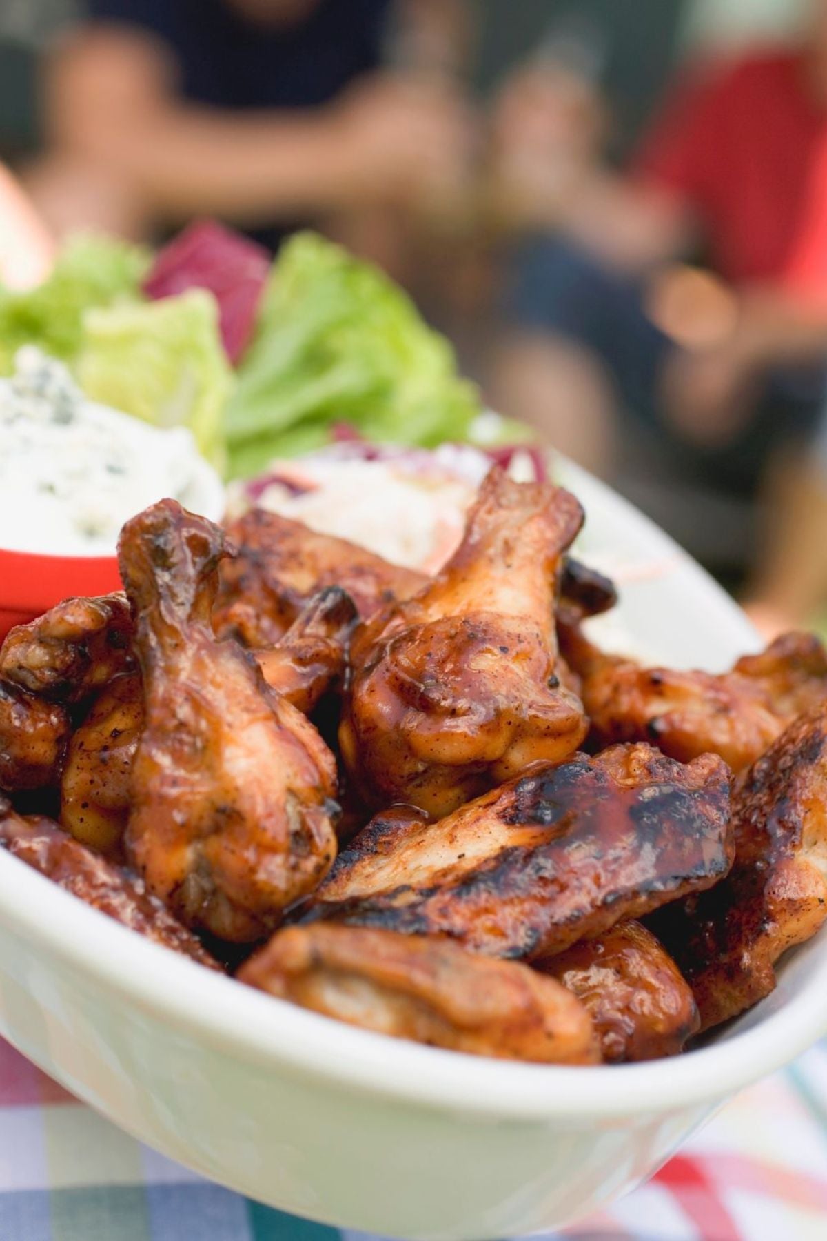 A white bowl filled with Grilled Chicken Wings sits on a table, accompanied by lettuce, dip, and other salad ingredients in the background.
