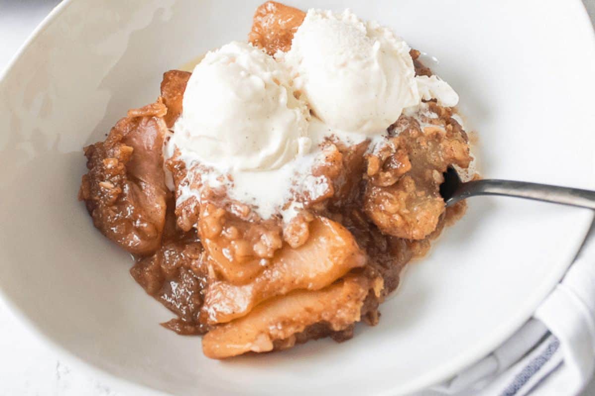 A bowl of apple cobbler, one of those classic desserts, topped with two scoops of vanilla ice cream and a spoon on the side.
