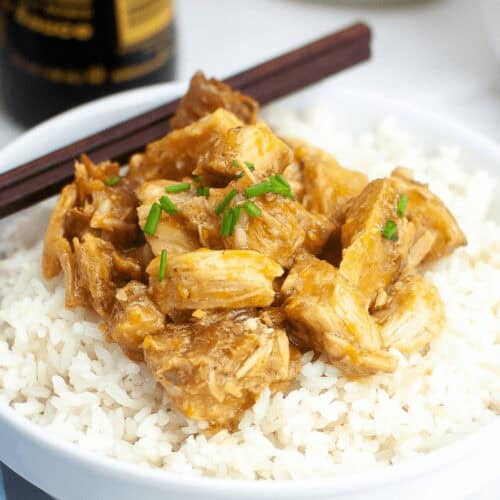 A white bowl contains fluffy white rice topped with chunks of crockpot orange chicken, coated in a savory sauce and garnished with chopped green herbs. Chopsticks rest on the rim of the bowl.