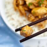 Close-up of chopsticks holding a piece of crockpot orange chicken over a bowl of rice topped with more meat. A blue cloth is partially visible on the left side of the image.