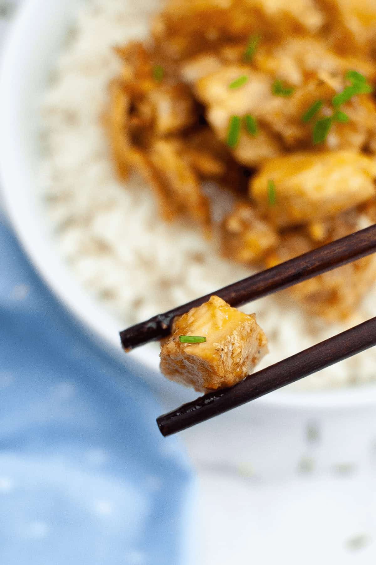 Close-up of chopsticks holding a piece of crockpot orange chicken over a bowl of rice topped with more meat. A blue cloth is partially visible on the left side of the image.