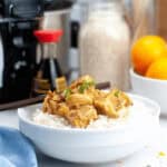 A white bowl filled with rice and topped with crockpot orange chicken garnished with green herbs is placed on a kitchen counter with various kitchen items and ingredients visible in the background.