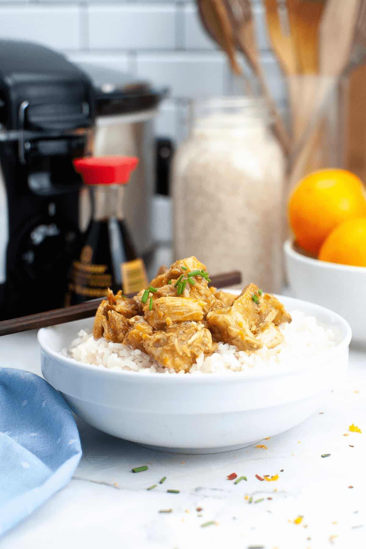 A white bowl filled with rice and topped with crockpot orange chicken garnished with green herbs is placed on a kitchen counter with various kitchen items and ingredients visible in the background.