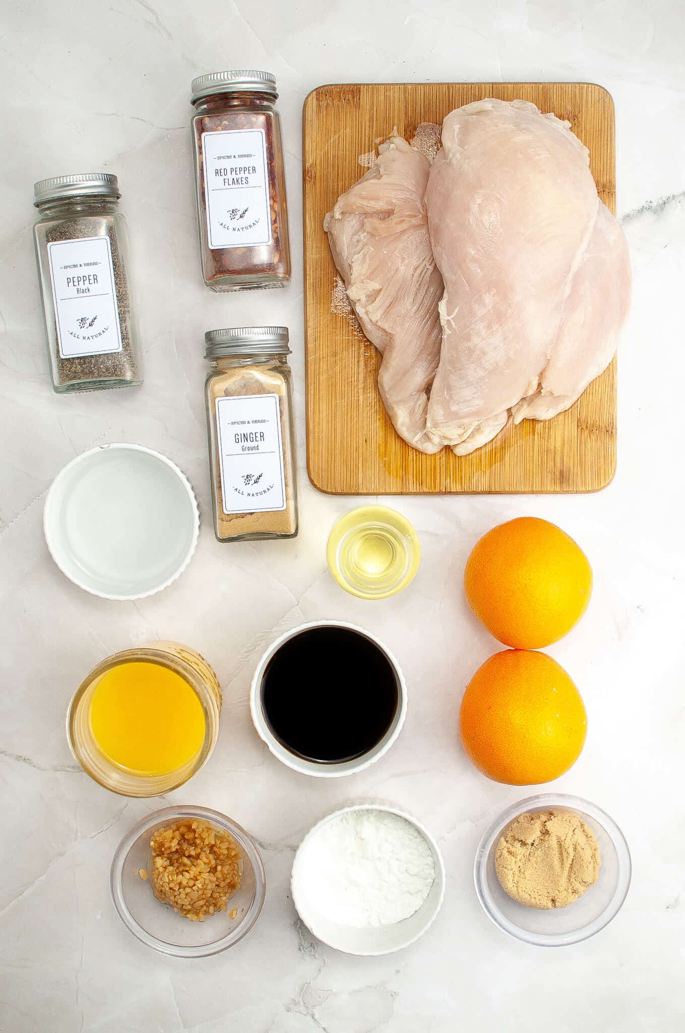Top-down view of ingredients for a recipe, featuring raw chicken breasts, seasonings, orange, orange juice, soy sauce, minced garlic, cornstarch, and brown sugar on a white surface.