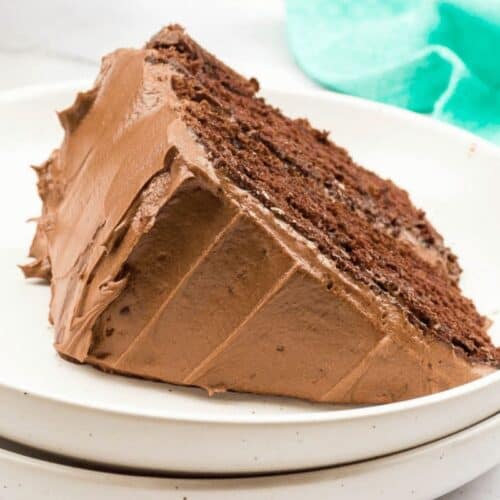 A slice of chocolate cake with rich chocolate frosting on a white plate. The cake has two layers and a moist texture, presented on a simple, clean backdrop with a green cloth visible—truly one of those old-fashioned dessert recipes that never go out of style.