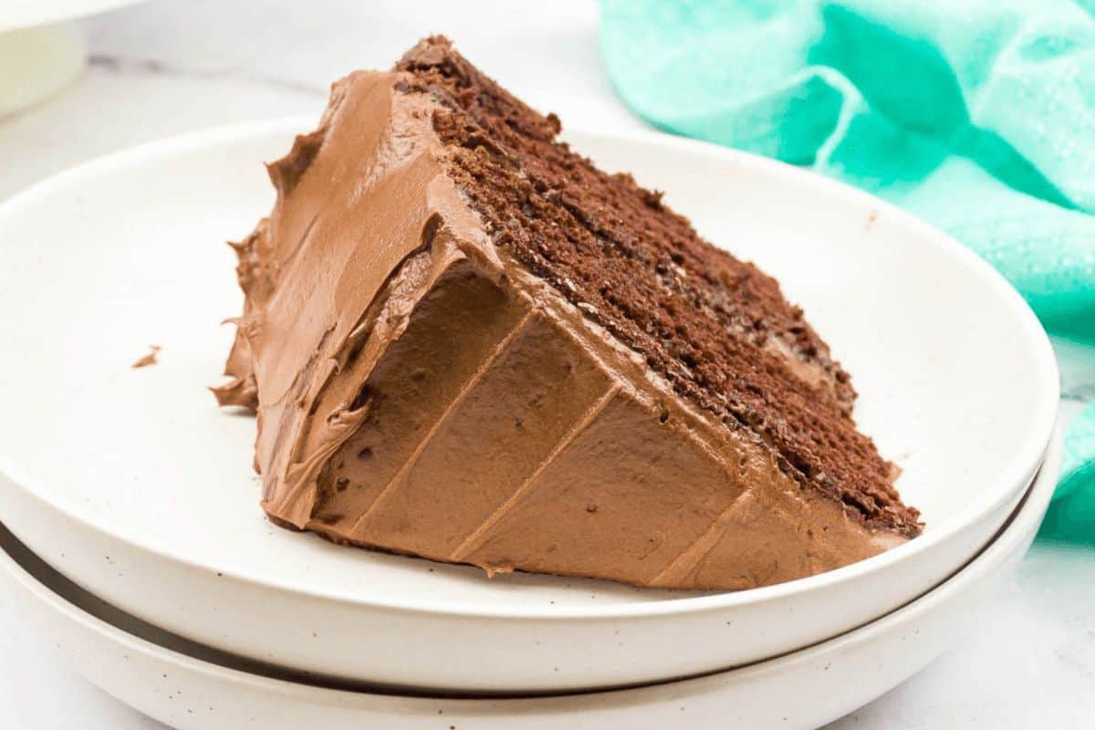 A slice of chocolate cake with rich chocolate frosting on a white plate. The cake has two layers and a moist texture, presented on a simple, clean backdrop with a green cloth visible—truly one of those old-fashioned dessert recipes that never go out of style.