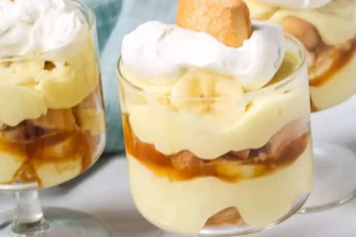 Two glasses of banana pudding topped with whipped cream and a vanilla wafer on a white surface, with a blurred blue cloth in the background. This old-fashioned dessert recipe features layers of bananas, custard, and wafers that never go out of style.