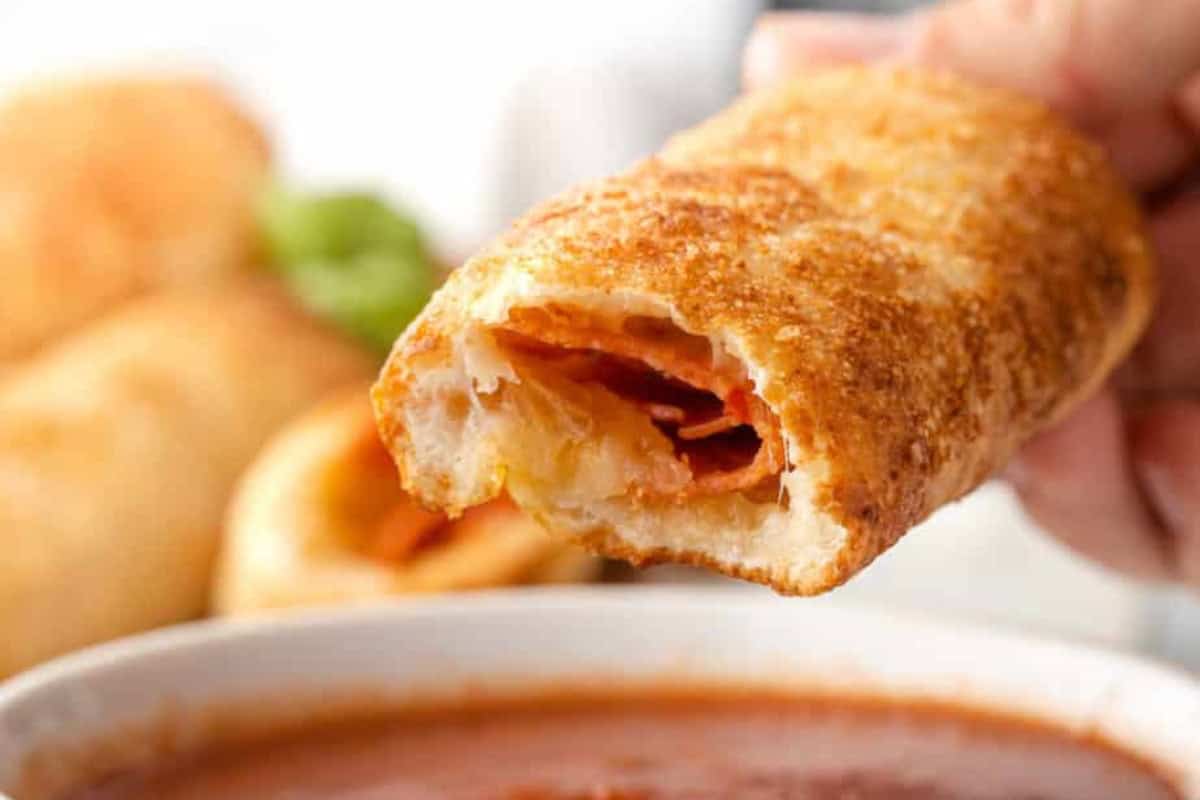 A hand holding a piece of pepperoni roll with a bite taken out of it hovers over a ramakin of marinara sauce.