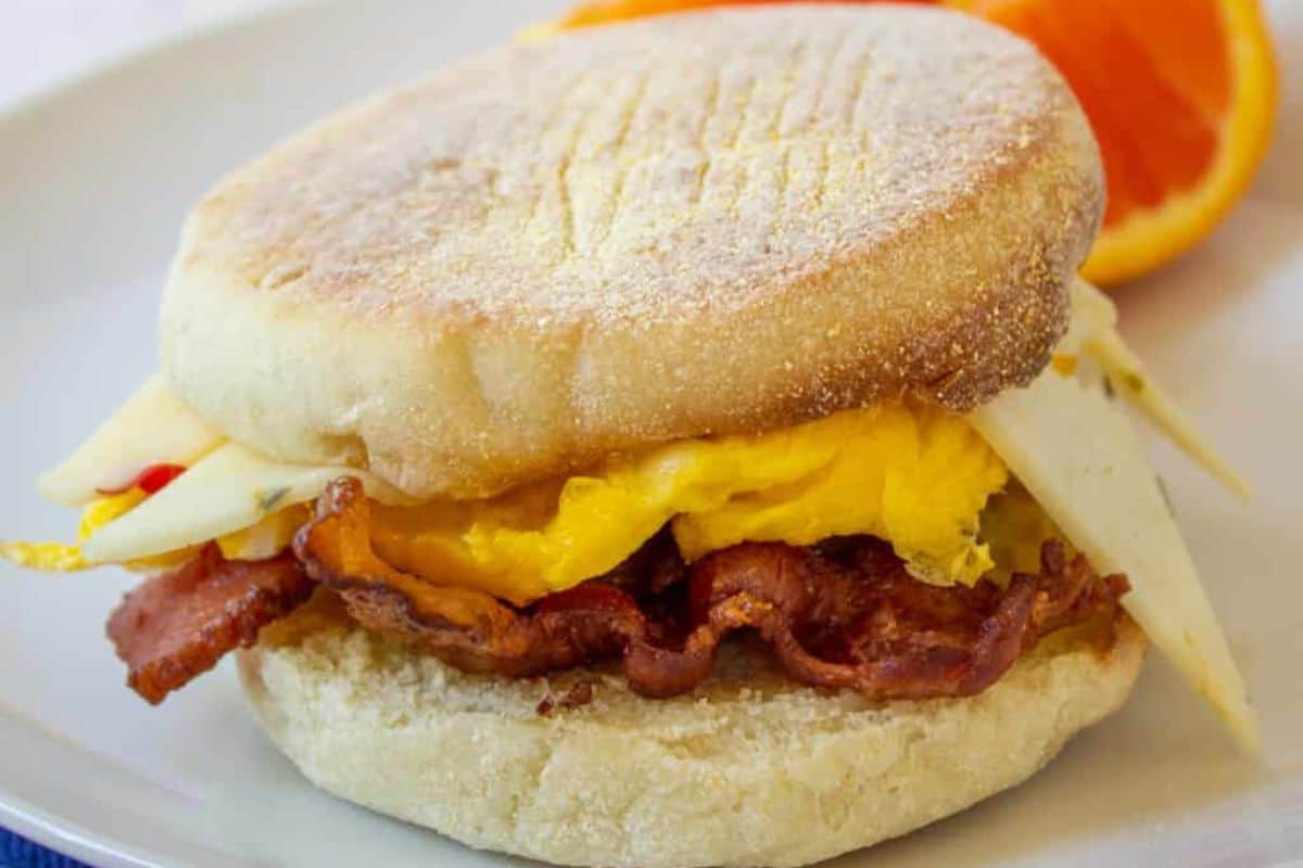 A close-up of an English muffin breakfast sandwich with bacon, egg, and cheese on a white plate.