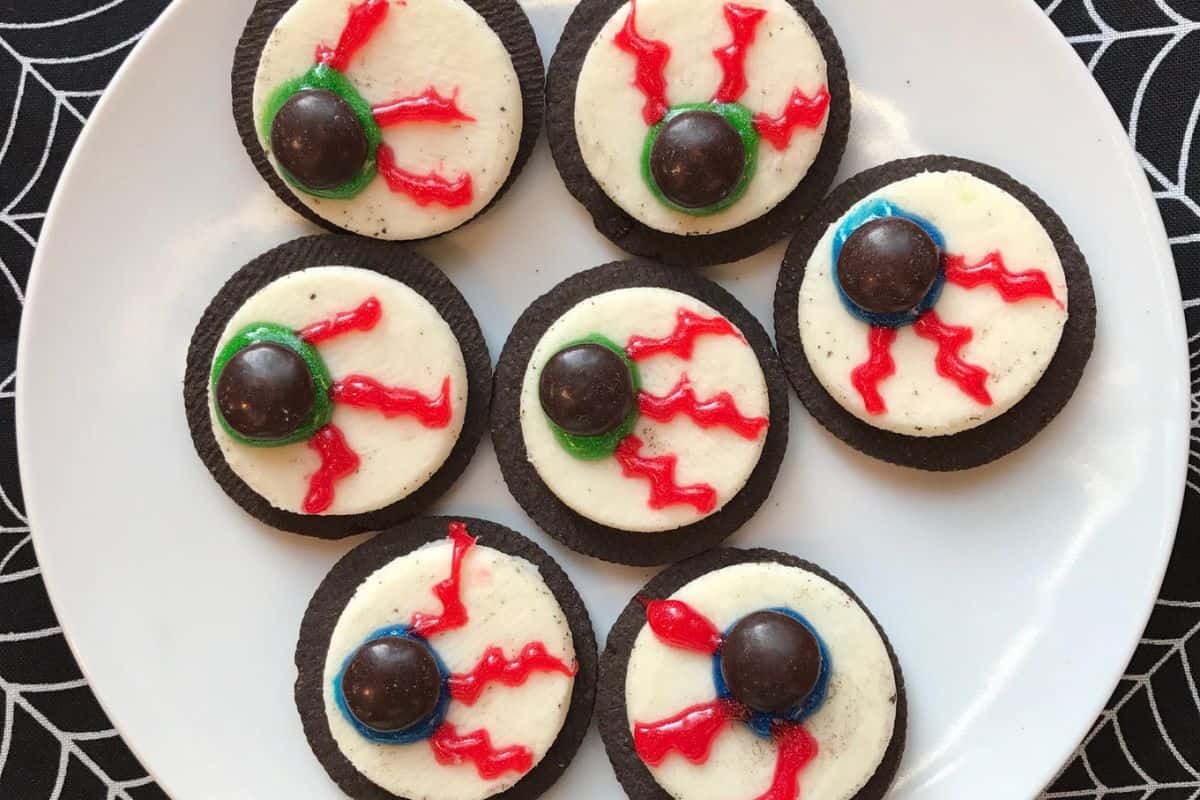 A white plate holding seven Halloween-themed cookies decorated to look like eyeballs with cream, chocolate, and red icing for veins. The plate is on a table with a black spiderweb-patterned cloth, perfect for your Scream Desserts collection.