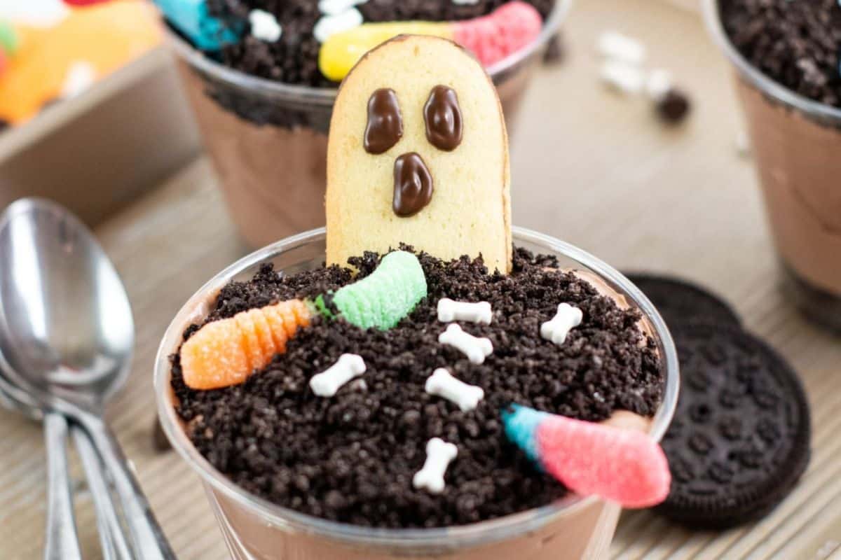 A scream dessert cup with crushed cookies resembling dirt, gummy worms, candy bones, and a ghost-shaped cookie sits on top. Spoons and cookies are visible in the background. Perfect for your collection of Halloween recipes!
