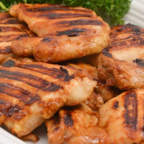 Asian Grilled Chicken thighs with visible char marks are arranged on a white plate, garnished with a bunch of fresh parsley in the background.