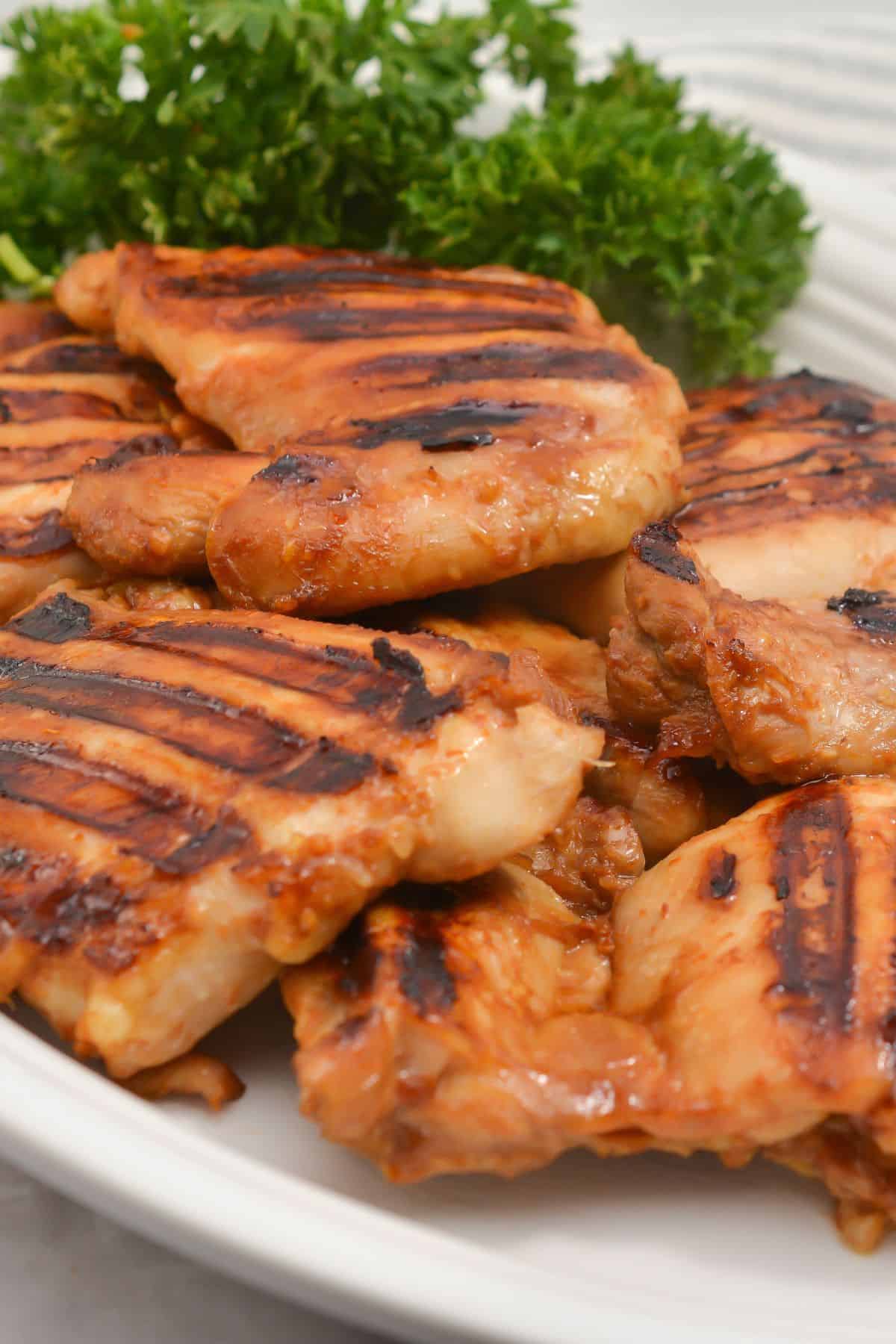 Asian Grilled Chicken thighs with visible char marks are arranged on a white plate, garnished with a bunch of fresh parsley in the background.