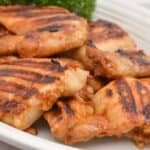 Asian Grilled Chicken thighs with visible char marks are served on a white plate with lemon wedges and greens on the side.