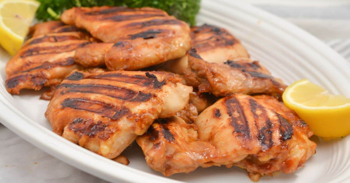 Asian Grilled Chicken thighs with visible char marks are served on a white plate with lemon wedges and greens on the side.