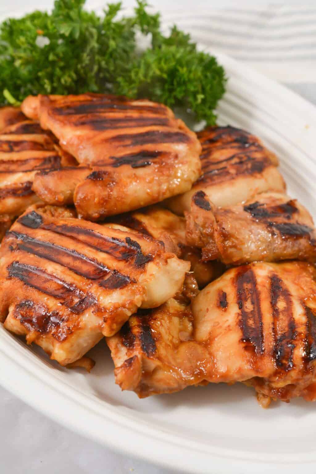 Asian Grilled Chicken thighs with visible char marks are arranged on a white plate, topped with fresh parsley garnish in the background.