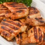 Asian Grilled Chicken thighs with visible char marks are arranged on a white plate, topped with fresh parsley garnish in the background.