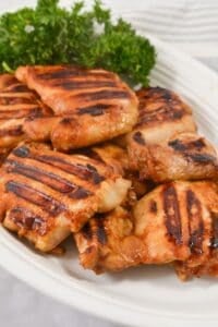 Asian Grilled Chicken thighs with visible char marks are arranged on a white plate, topped with fresh parsley garnish in the background.