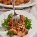 A hand uses a fork and knife to cut into Asian Grilled Chicken served on a bed of parsley, with a plate of more grilled chicken and a lemon wedge in the background.