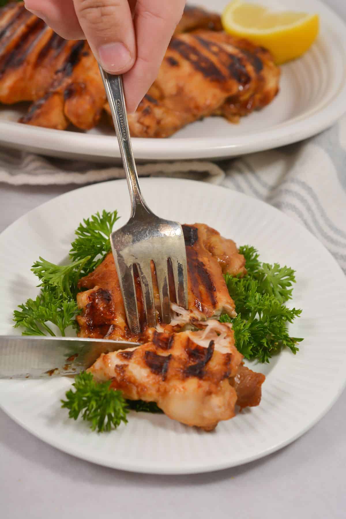 A hand uses a fork and knife to cut into the dish served on a bed of parsley, with a plate of more meat and a lemon wedge in the background.