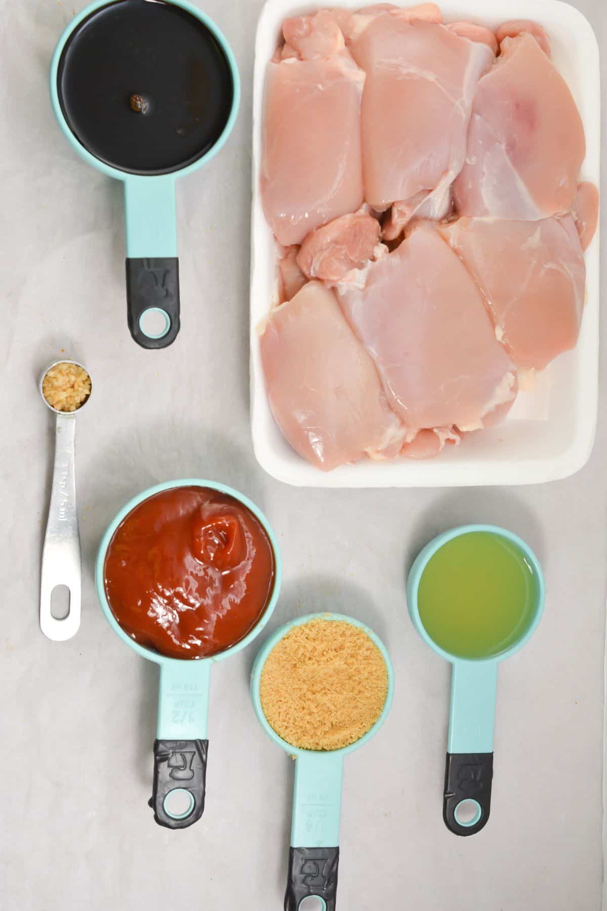 Raw meat in a tray, surrounded by measuring cups containing soy sauce, ketchup, bread crumbs, olive oil, and a teaspoon of minced garlic on a white surface.
