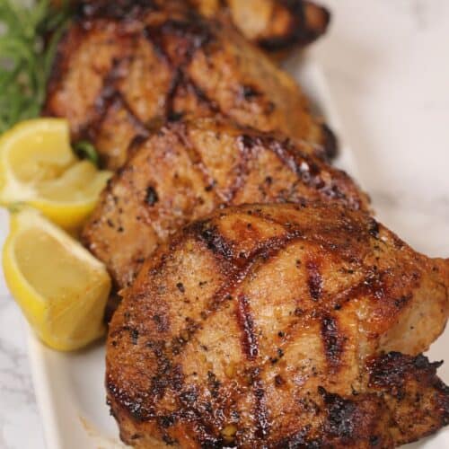 Four grilled pork chops, marinated in a savory grilled pork chops marinade and marked with perfect grill lines, are arranged on a rectangular white plate, garnished with lemon wedges and herbs on a marble surface.