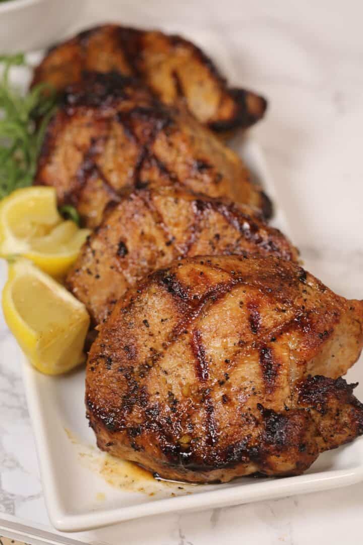 Four grilled pork chops, marinated in a savory grilled pork chops marinade and marked with perfect grill lines, are arranged on a rectangular white plate, garnished with lemon wedges and herbs on a marble surface.