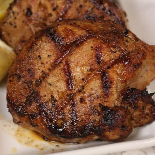 Grilled, seasoned chicken thighs with char marks on a white plate, garnished with lemon wedges—perfectly mouthwatering and just as flavorful as any Grilled Pork Chops Marinade.