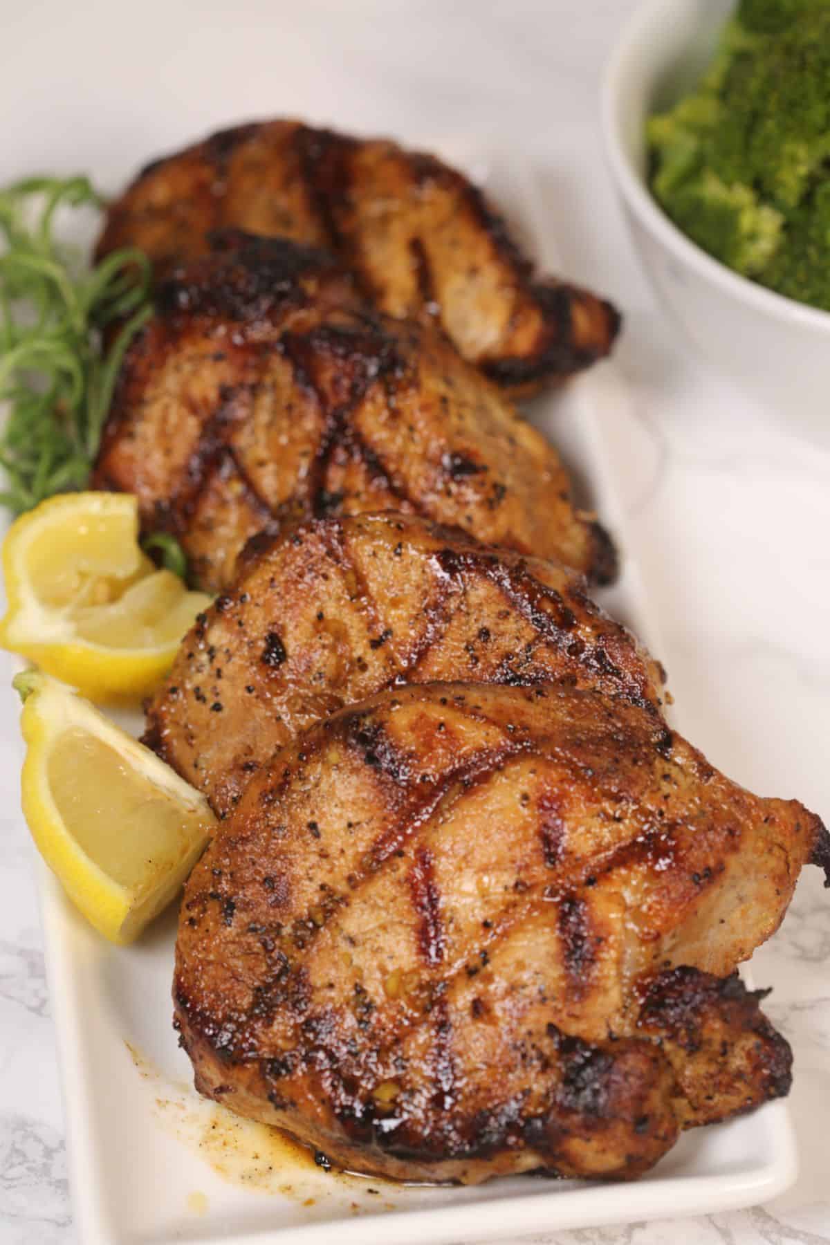 Four grilled pork chops, infused with a savory grilled pork chops marinade, display perfect grill marks on a rectangular white plate. They’re garnished with lemon wedges and fresh herbs, while a bowl of broccoli sits in the background for a wholesome touch.