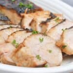 Sliced marinated pork loin, grilled to perfection and garnished with chopped parsley, served on a white oval plate.