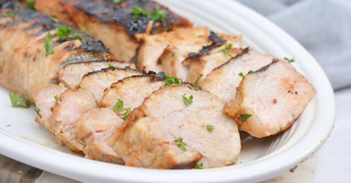 Sliced marinated pork loin, grilled to perfection and garnished with chopped parsley, served on a white oval plate.