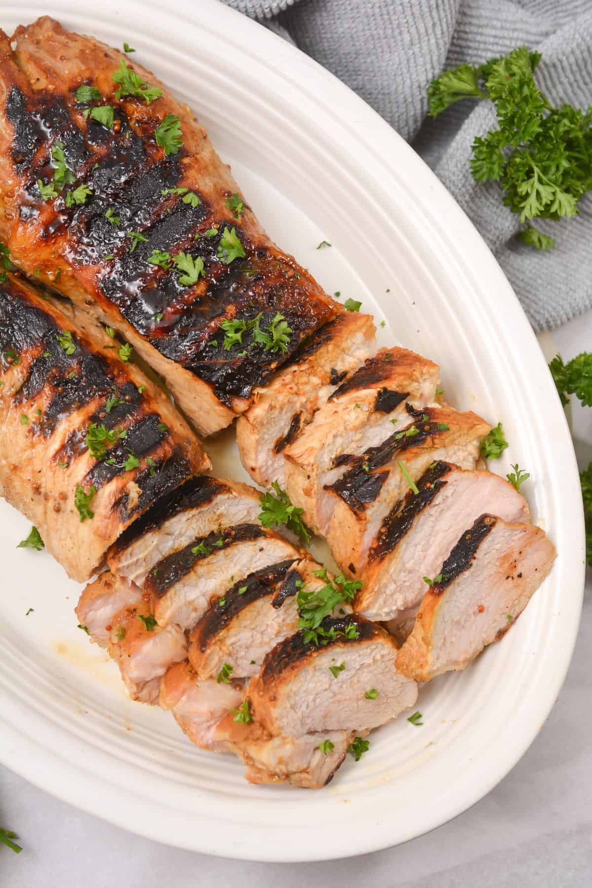 Tenderloin, partially sliced, garnished with chopped parsley, served on an oval white plate with a gray cloth and fresh parsley nearby.