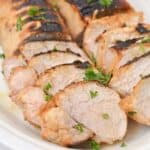 Sliced grilled marinated pork loin on a white plate, garnished with chopped parsley.