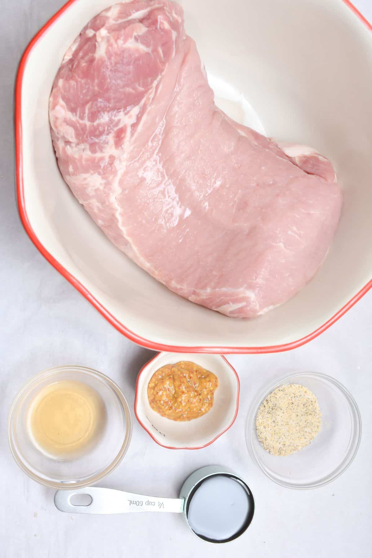 Raw meat in a bowl, surrounded by small bowls containing mustard, seasoned mustard, breadcrumbs, and a measuring cup of milk on a white surface.