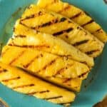 Grilled Pineapple Slices are arranged on a turquoise plate, their char marks visible against the golden fruit, set atop a striped cloth and woven surface.
