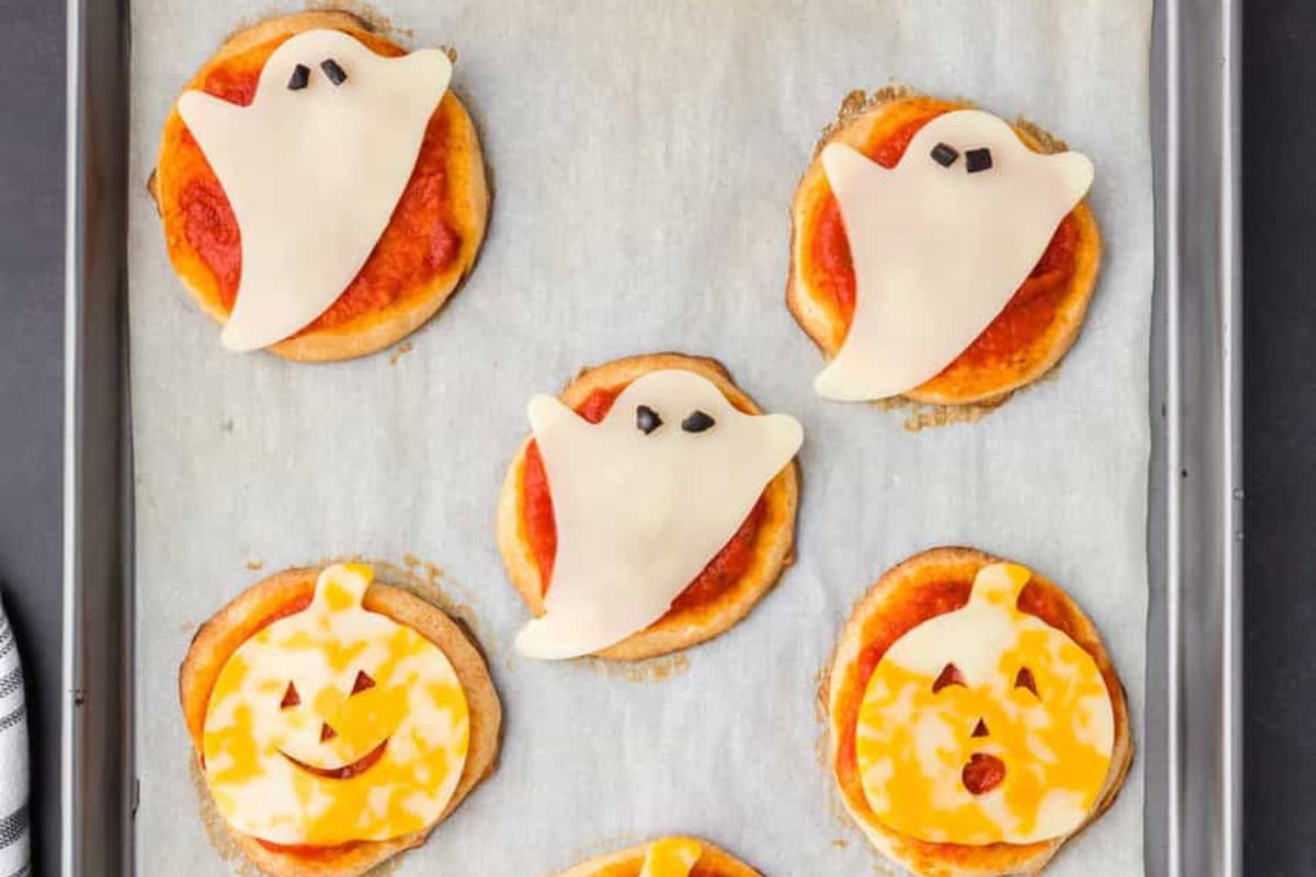 Five mini Halloween pizzas on a parchment paper lined baking sheet. The top two and middle pizzas have white cheese shaped like ghosts on top along with black olive bits for eyes, while the bottom two have jack o lanterns made out of Colby jack cheese.