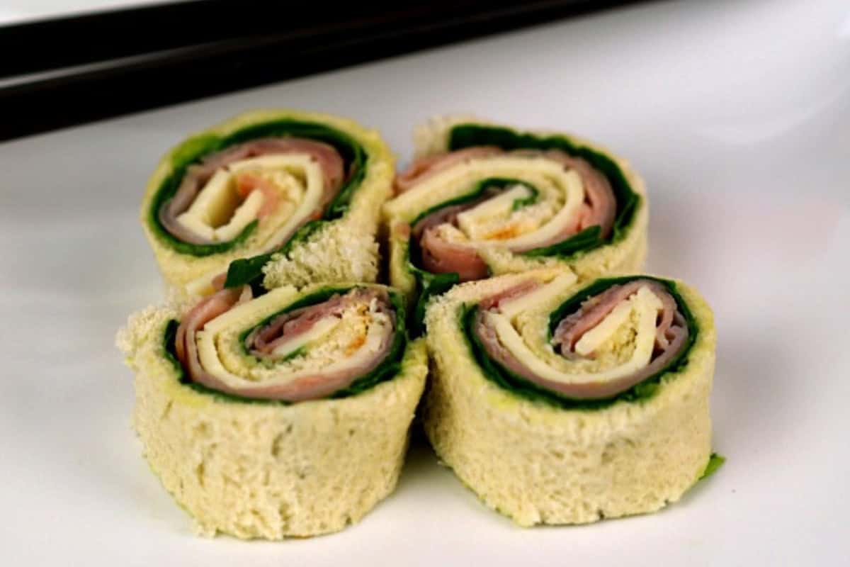 Four sandwich pinwheel "sushi" rolls with layers of bread, ham, cheese, and spinach are arranged on a white plate.