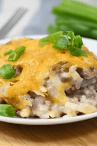 Close-up of a plate with a cheesy hamburger and hash brown casserole topped with melted cheese and green onions, with green onions in the background on a wooden surface.