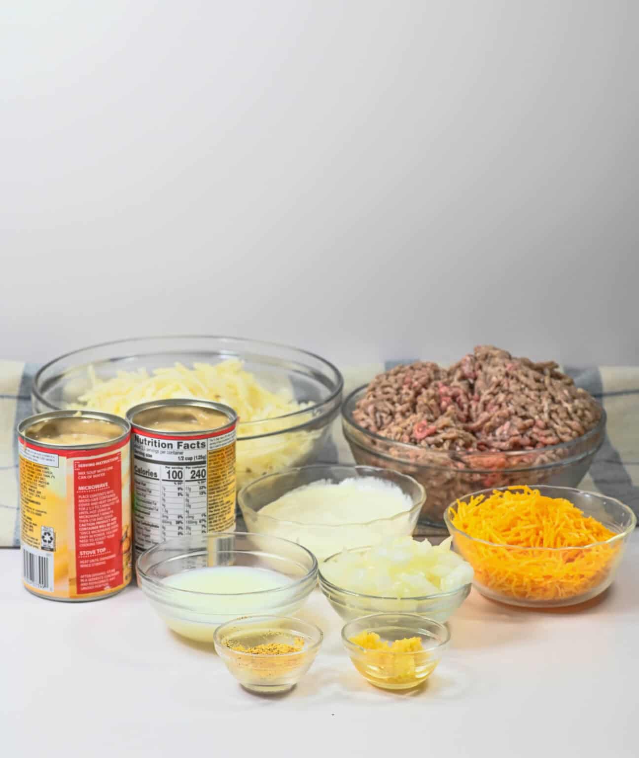 The table is set with ingredients for a delightful Hamburger and Hash Brown Casserole: canned tomatoes and cream, ground meat, shredded cheese, chopped onions, and small bowls brimming with spices, ready to create a comforting dish.