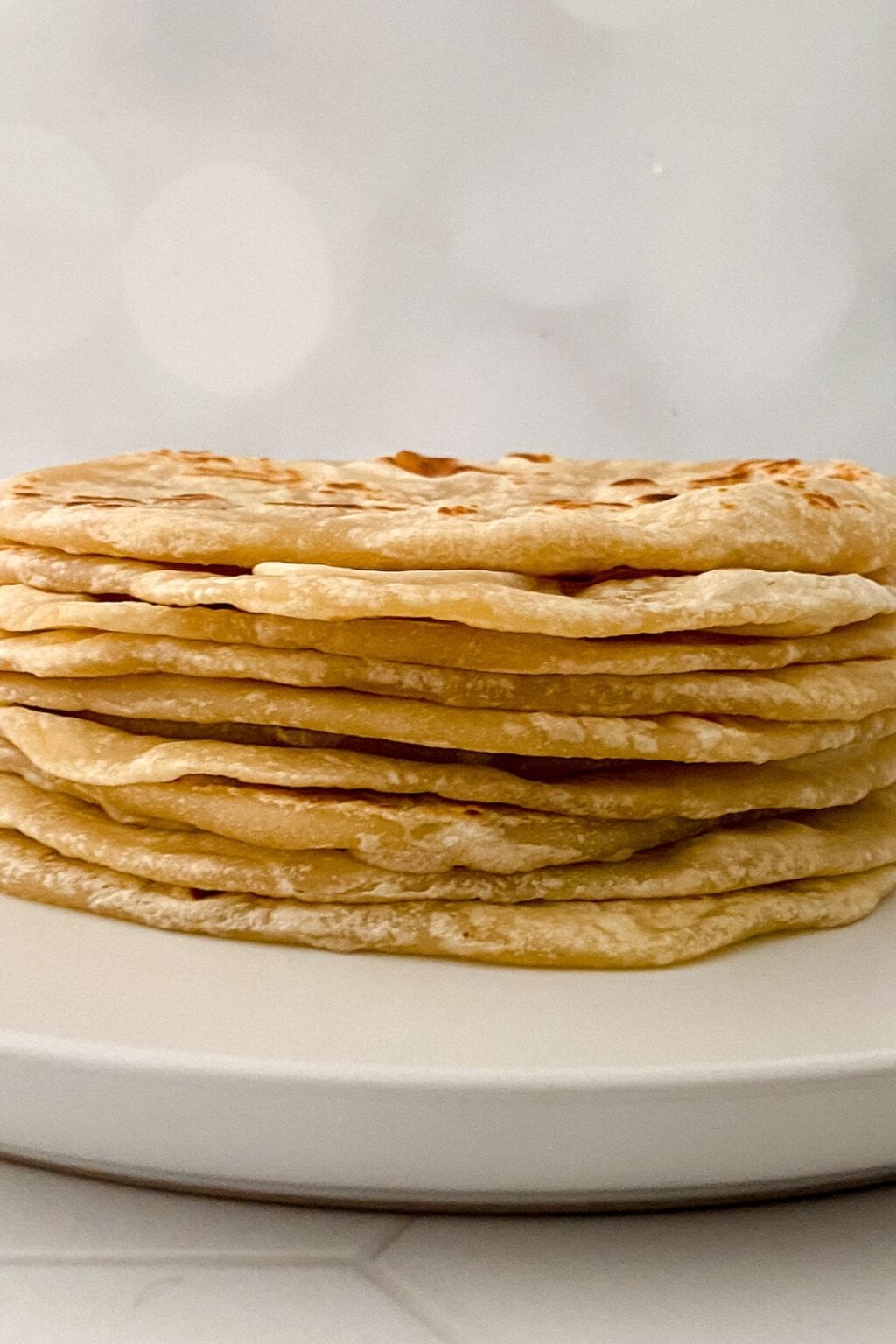 A stack of thin, golden-brown Homemade Flour Tortillas is arranged neatly on a white plate against a blurred, light-colored background.