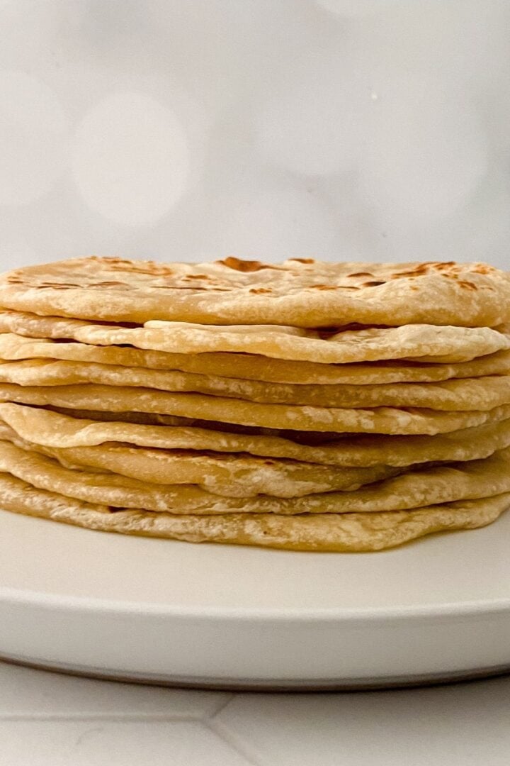 A stack of thin, golden-brown Homemade Flour Tortillas is arranged neatly on a white plate against a blurred, light-colored background.