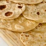 Several pieces of homemade flour tortillas, lightly browned, are stacked on a wooden cutting board.