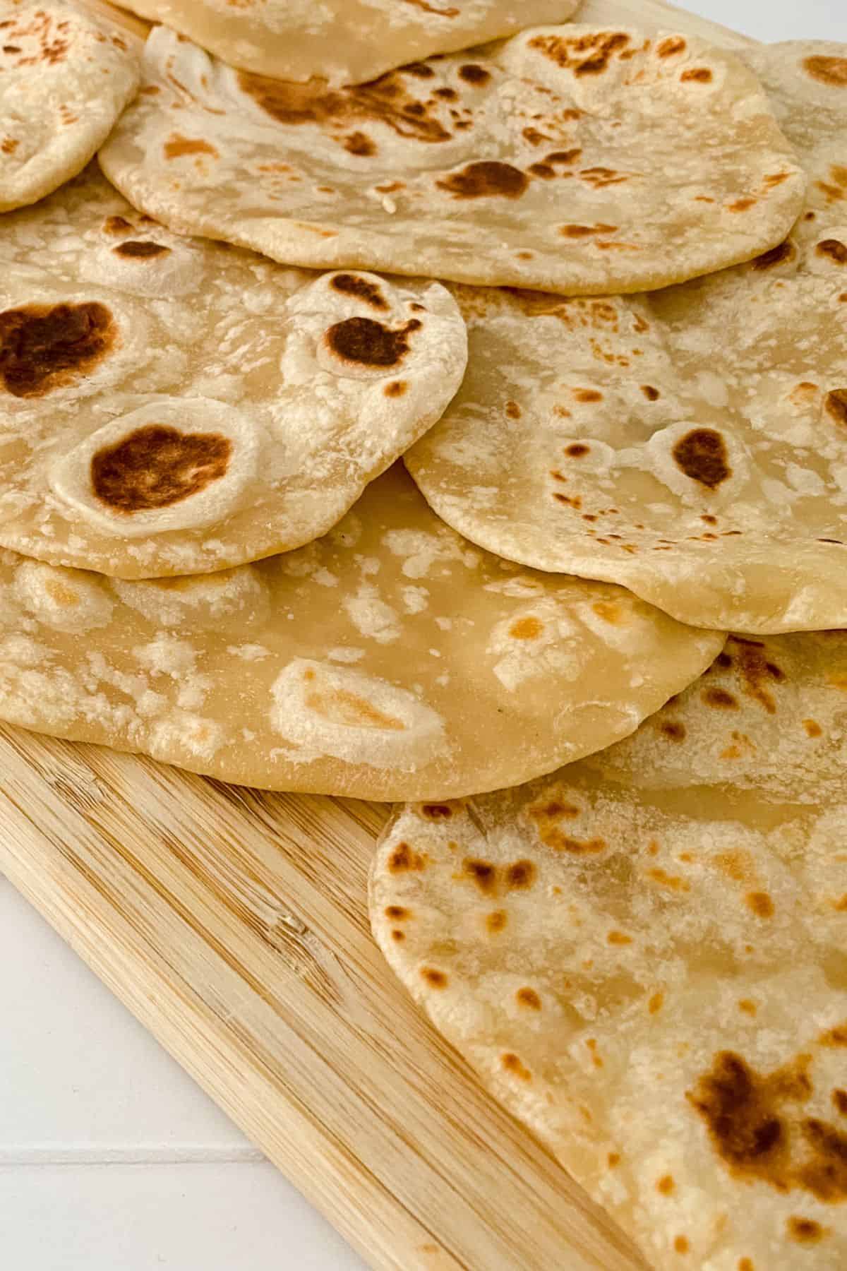 Several pieces of homemade flour tortillas, lightly browned, are stacked on a wooden cutting board.