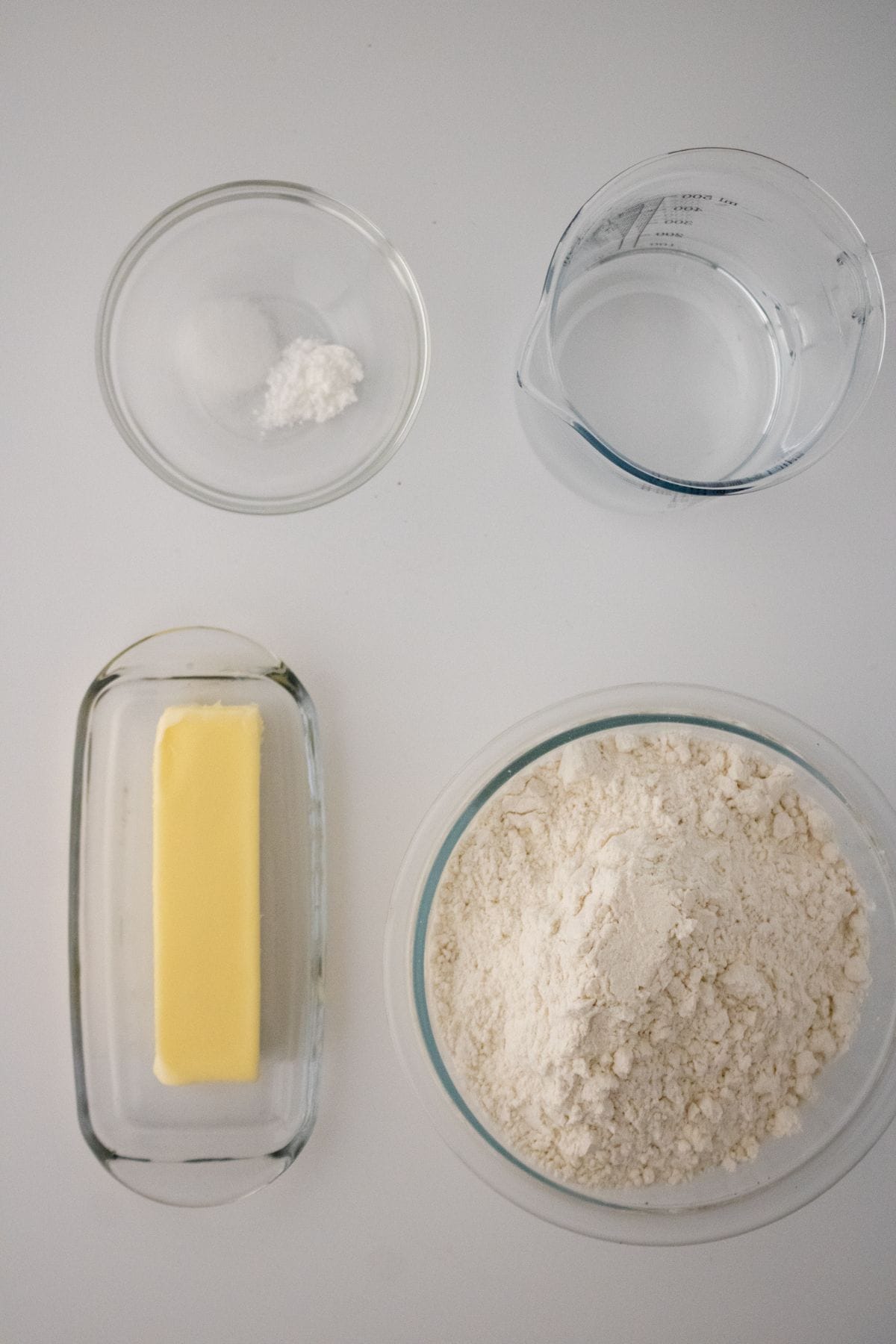 Ingredients for Homemade Flour Tortillas—butter, flour, water, salt, and baking powder—are arranged in separate containers on a white surface.