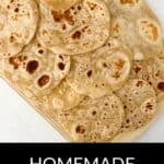 A wooden board holds several round, lightly browned homemade flour tortillas on a white surface. Text below reads "Homemade Flour Tortillas.