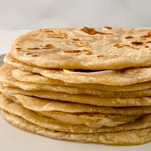A stack of homemade flour tortillas is arranged on a white plate against a plain background.